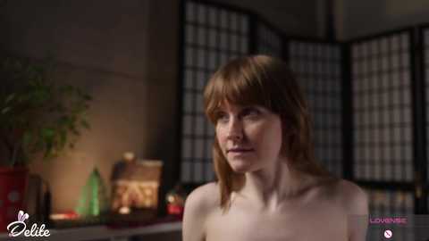 Media: Video of a topless, fair-skinned woman with straight, light brown hair, standing indoors near a potted plant and Japanese folding screen.