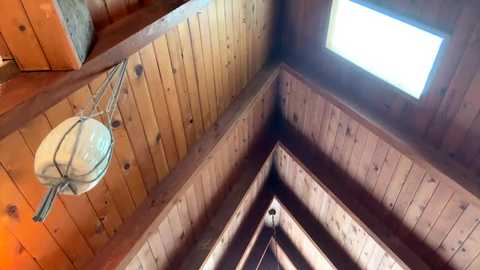 Video of a wooden room with a triangular ceiling and a small skylight. The walls and ceiling are made of light brown, vertically aligned planks, creating a rustic, cabin-like atmosphere. A metal lamp hangs from the ceiling.
