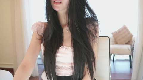 A video of a young woman with long, straight black hair, fair skin, and a slender build, wearing a pale pink lace top, seated indoors, with a beige chair and light-colored walls in the background.