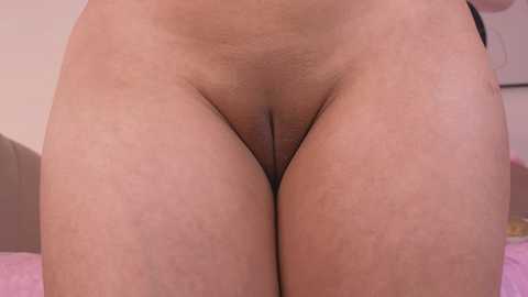 Video of a close-up, nude, light-skinned female torso, showing her pubic area and slightly visible labia, with a blurred background of pink and white bedding.