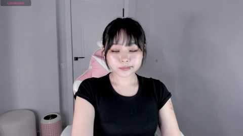 Video of a young Asian woman with pale skin, straight black hair, and a black t-shirt, sitting with eyes closed and mouth open, against a gray wall.