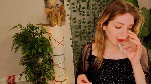Video of a fair-skinned, blonde woman in a black sequined top, sipping water. Background includes a white vase with gold and red ribbons, green plants, and a string of green leaves.