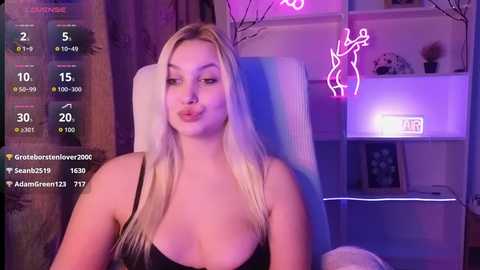 Video of a blonde, fair-skinned woman in a black tank top, seated on a white chair, with a digital screen displaying her social media metrics in the background.