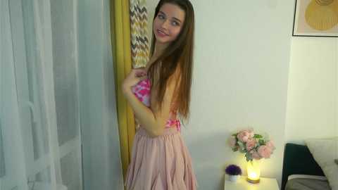 Video of a young, slender woman with long brown hair, wearing a pink floral top and a light pink pleated skirt, standing in a modern, brightly lit bedroom with pastel-colored decor and a flower arrangement on a nightstand.