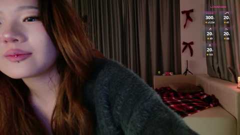 A video shows a young Asian woman with long, straight brown hair, wearing a gray hoodie. She has a nose piercing and pink lipstick. Background features a cozy living room with a red plaid blanket and holiday decorations.