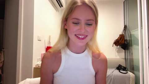 Video of a smiling, blonde-haired young woman in a white tank top, standing in a modern, minimalist bedroom with an air conditioner, a bed, and hanging bags.