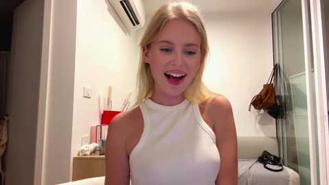 Video of a smiling, blonde woman with fair skin, wearing a white sleeveless top, standing in a modern bedroom with white walls and a glass door.