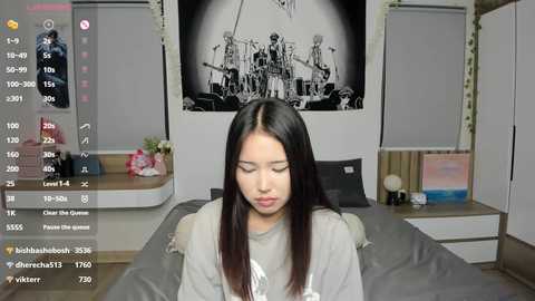 Video of a young East Asian woman with straight black hair, wearing a grey sweater, sitting on a bed with a grey blanket, in a modern bedroom with a black-and-white poster of a warship on the wall.