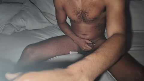 Video of a nude, dark-skinned man with a muscular build, sitting on a bed with white sheets. He holds a white vibrator against his penis.