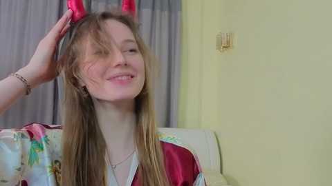 Video of a young, smiling Caucasian girl with long, light brown hair, wearing red devil horns, adjusting them with a hand. She is in a yellow-walled room with a white bed and a pink and green bedspread.