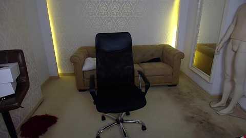 Video of a dimly lit room with a beige sofa, a black office chair, and a mannequin dressed in a white gown standing near a mirror. The walls have intricate white wallpaper and yellow accent lighting.