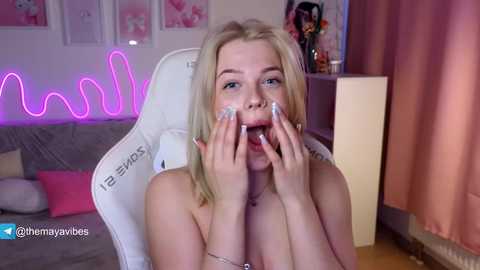 Video of a blonde, fair-skinned woman with light makeup, covering her mouth with hands, in a pink-lit, cozy room with a white gaming chair, pink cushions, and a neon \"molly\" sign.