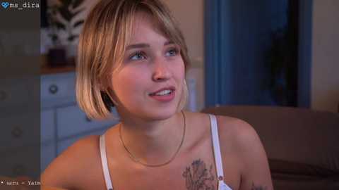 Video of a young woman with short blonde hair and fair skin, wearing a white tank top, smiling in a dimly lit room with a dresser and potted plant in the background.