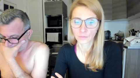 Media: Video of a middle-aged man with grey hair and glasses, shirtless, and a woman with long blonde hair and glasses in a kitchen.
