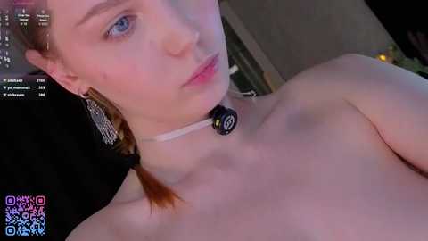 Media: Video of a pale-skinned, red-haired young woman with blue eyes, wearing a black choker with a white ribbon and silver earrings. She is topless, with her breasts partially visible. The background is dimly lit, with a blurred, dark-colored wall.