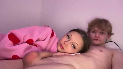 Media: Video of a nude young man and woman lying close in bed, covered with a pink blanket. The woman has long brown hair, and the man has curly blond hair.