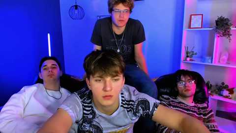 Video of four teenage boys in a dimly lit room with blue and purple lighting. One boy sits on a chair, others sit on a couch, wearing casual clothing and glasses.