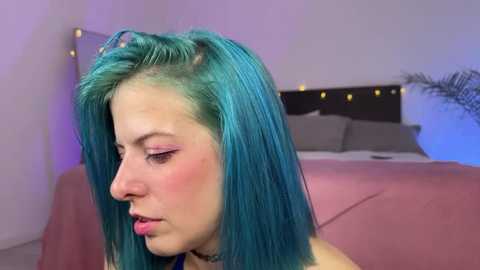 Video of a fair-skinned woman with vibrant teal hair, wearing a black choker, in a dimly lit bedroom with a gray bed and fairy lights.