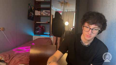 Video of a young man with short, curly hair, wearing glasses, black shirt, and standing near a bed with a pink blanket in a dimly lit room.