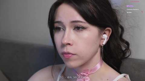 Video of a young woman with long dark hair, fair skin, wearing a pink choker and white earbuds, sitting on a gray couch, looking introspective.