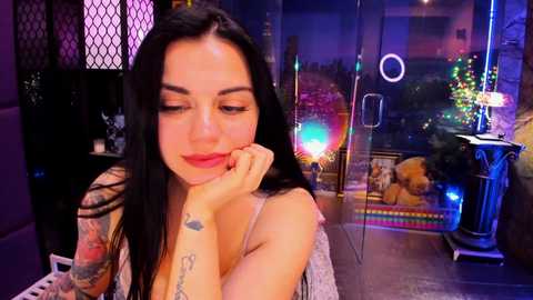 A video of a young woman with long dark hair, tattoos, and red lipstick, sitting indoors with a glass wall behind her.