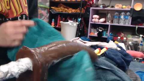 Video of a messy workshop with a person in the foreground holding a brown, wooden object covered in white powder. The background shows shelves filled with various items, including bottles and tools.