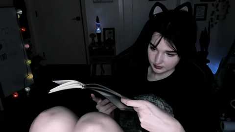 Video of a young woman with long black hair, wearing cat ears, reading a book in a dimly lit room with a mirror and decorative lights on the left.