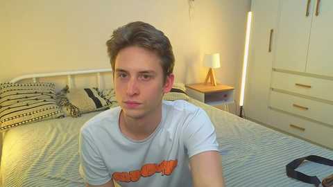 Video of a young man with short brown hair, wearing a white T-shirt with an orange graphic, sitting on a bed with striped bedding in a beige, minimalistic bedroom.