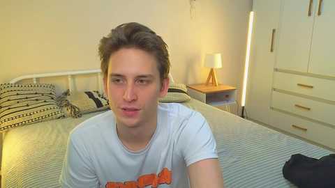 Video of a young, fair-skinned man with short brown hair, wearing a white t-shirt with an orange graphic, sitting on a bed in a modern, minimalist bedroom with a striped bedspread and white furniture.