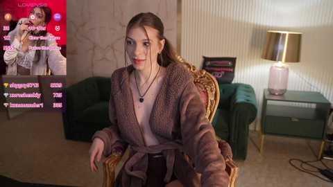 Video of a young woman with light skin, brown hair, and a beige cardigan, seated in a plush chair, wearing headphones. Background features a TV, green couch, and a lit lamp.