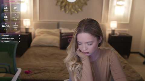 Video of a blonde woman in a brown top, looking down, in a cozy, dimly lit bedroom with beige walls, a bed, and two nightstands with lamps.