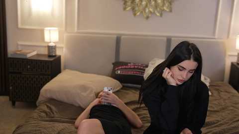 Video of a woman with long black hair, dressed in a black top, lying on a bed with beige bedding, talking on a phone, in a cozy bedroom with a white wall and decorative lamp.