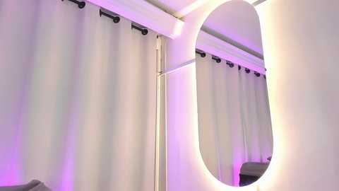 Video of a modern bathroom with a large oval mirror mounted on a white wall, reflecting the adjacent shower curtain. The curtain is light grey with black metal rings, and the room is illuminated by a soft purple light.
