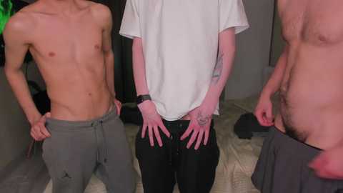 Video of three shirtless men in a dimly lit room; one in gray sweatpants, one in black pants, and the third partially undressed, revealing a tattooed arm.