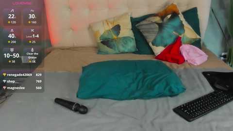 Video of a messy bedroom with a teal pillow, a black keyboard, a black microphone, and colorful pillows on a beige bed.