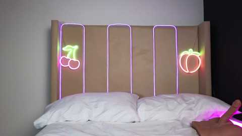 Video of a minimalist bedroom with a light beige headboard featuring neon pink and green fruit illustrations (cherries, peaches) against a plain white wall. The bed has white pillows and sheets, and a hand is visible adjusting the pillow.