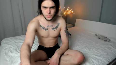 Video of a lean, shirtless man with long black hair and tattoos on his chest and arms, sitting cross-legged on a white bed, wearing black briefs, in a modern, dimly-lit bedroom.
