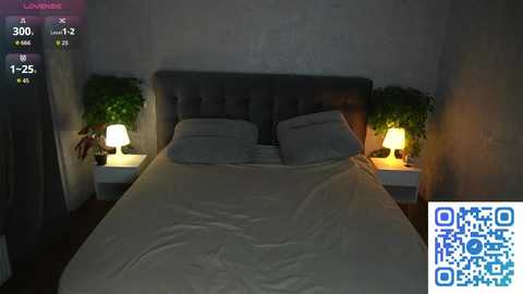 Video of a dimly-lit bedroom with a gray upholstered headboard, white pillows, and a cream-colored duvet. Nightstand lamps cast a warm glow. QR code overlay.
