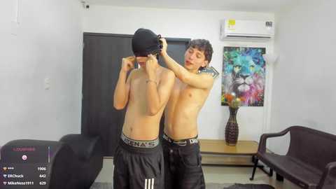 Video of two shirtless young men in black underwear, adjusting each other's hats in a minimalist room with colorful art on the wall.