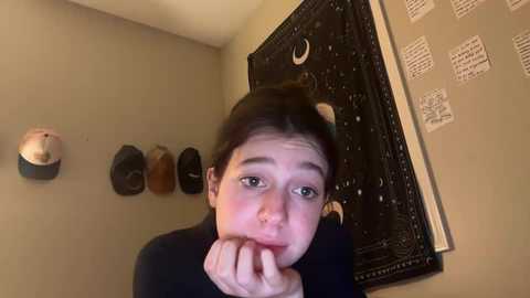 A video of a young woman with fair skin and dark hair in a messy bun, resting her chin on her hand, in a dimly lit room with beige walls and a black tapestry adorned with white musical notes.