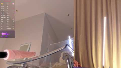 A video of a nude woman's right hand holding a realistic, erect, flesh-toned dildo, positioned beside a bed with beige curtains. The room has white walls and a modern, minimalist design.