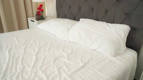 Video of a neatly made bed with white linens and a grey, tufted headboard. A bedside table holds a lamp and red flowers. Cream-colored curtains hang in the background.