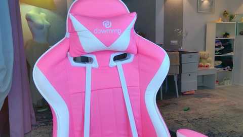 Video of a pink and white gaming chair with a sleek design, featuring a built-in headrest and adjustable armrests. Background includes a cluttered room with shelves, toys, and a plush teddy bear.
