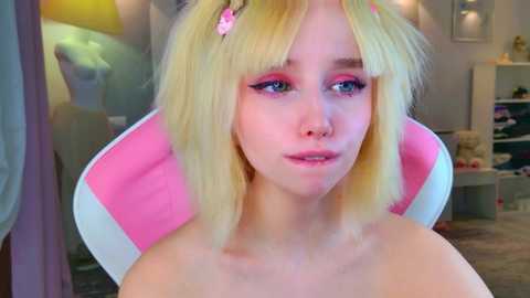 Video of a young, fair-skinned woman with blonde hair, styled in a bob with pink flowers, wearing pink and white headphones, sitting in a pink chair, wearing a light pink top, in a brightly lit room.