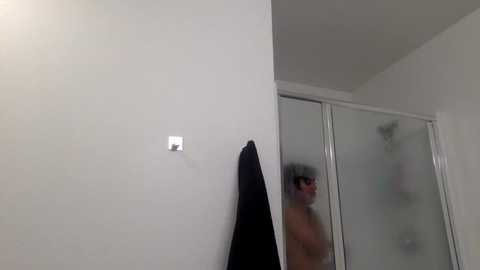 A video of a man with short, dark hair and glasses, partially obscured by steam, standing shirtless in a white-tiled bathroom with a black towel hanging on a wall and a glass shower door.