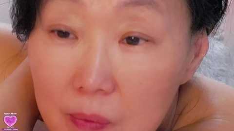 A close-up video of an East Asian woman with fair skin, short black hair, and light makeup, lying on her stomach, with a blurred background.