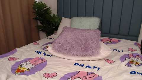 Media: Video of a cozy bedroom with a bed covered in a cartoon-themed comforter featuring Disney's \"Dumbo.\" A plush, lavender pillow rests on a white pillow, both on a grey upholstered headboard.