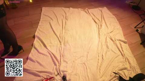 A video shows a beige, textured blanket with creases and folds, laid on a wooden floor, partially covering a black high heel shoe. The setting appears to be a dimly lit room, with a QR code in the bottom left corner.