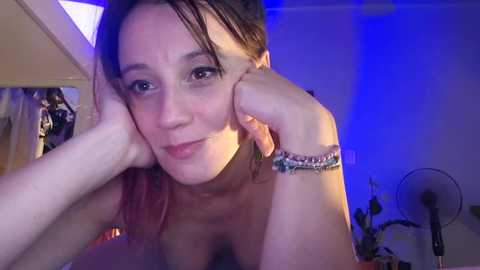 A video of a topless woman with fair skin, brown hair, and a light blue bracelet, leaning forward with her elbows on a bed, under blue lighting, in a bedroom.