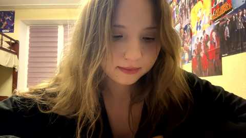 Video of a young, light-skinned girl with long, wavy brown hair, wearing a black jacket, looking down thoughtfully in a brightly decorated bedroom with posters and a bunk bed.